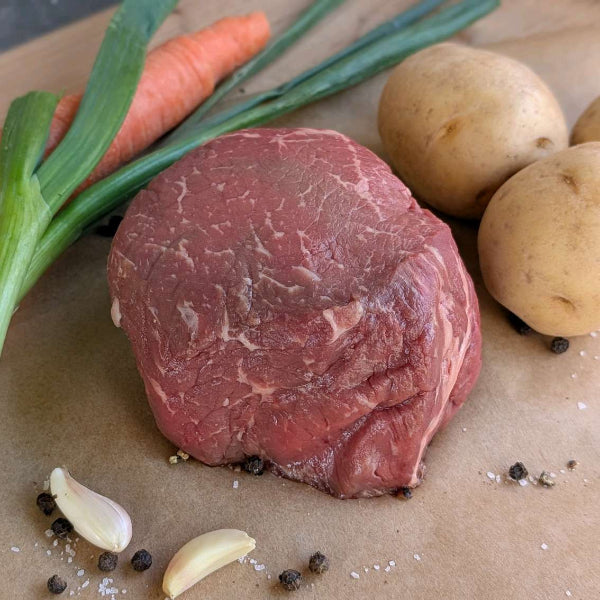 Top Sirloin sits on parchment paper with a carrot, green onions, potatoes, garlic cloves, and black pepper scattered around it.
