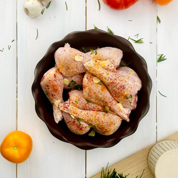 Raw chicken legs seasoned with herbs and spices, presented in a dark brown scalloped bowl on a white wooden table, surrounded by garlic, rosemary, and citrus slices.