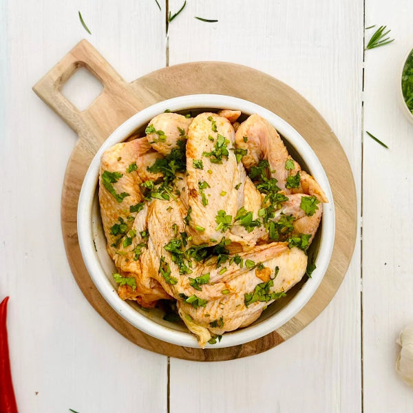 Marinated chicken wings garnished with chopped parsley, served in a white bowl on a round wooden board, set on a white wooden table with red chili peppers, garlic, and a small bowl of parsley.