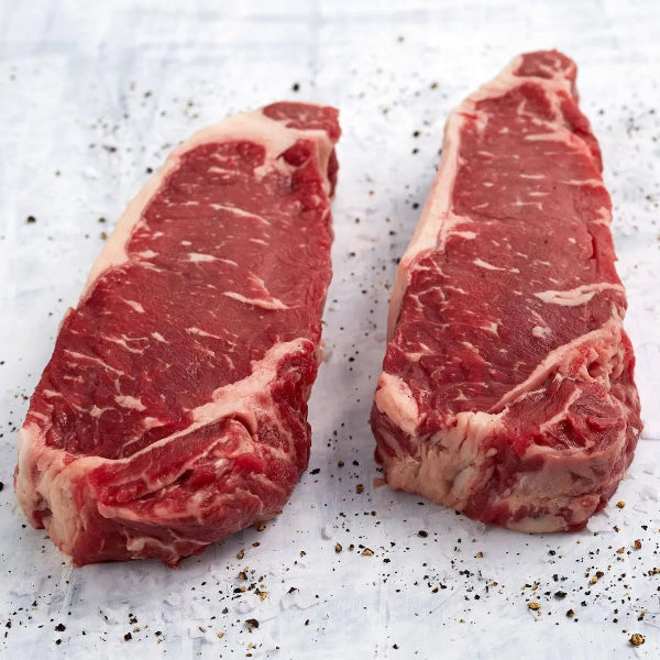 Two NY Strip Steaks on a white surface with black pepper sprinkled around.